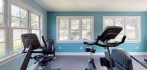 This single-floor fitness center offers a clean, energizing environment designed for effective workouts. The space is finished with soft teal walls and crisp white trim, creating a refreshing and motivating atmosphere. Durable rubber tile flooring covers the workout area, providing both safety and comfort underfoot.

The facility is generously equipped with a variety of cardio machines, including treadmills, an elliptical trainer, a recumbent bike, and an upright stationary bike, accommodating a range of fitness preferences. A large wall-mounted mirror allows users to monitor their form, while exercise mats are available for stretching and floor work. A wall-mounted television keeps exercisers entertained during their sessions.

Natural light pours in through multiple white-trimmed windows along multiple walls, offering pleasant views of the surrounding greenery outdoors. Overhead fluorescent lighting ensures the space remains well-lit at all times. A dedicated entry door with a glass panel provides convenient direct outdoor access. Additional amenities include a bulletin board with posted fitness center rules and information, a water station, and a paper towel dispenser for maintaining hygiene. The overall layout is thoughtfully organized to maximize open floor space while keeping all equipment easily accessible.