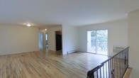 This single-floor residence offers a clean, open layout designed for comfortable everyday living. The main living area is anchored by light-toned hardwood flooring that extends throughout, lending warmth and continuity to the space. A large sliding glass door floods the room with natural light and provides direct access to an outdoor deck, where lush greenery creates a serene backdrop. A baseboard heating unit runs along the wall, ensuring year-round comfort. The open flow connects the living area to the kitchen, which features dark wood cabinetry and a black refrigerator, offering a subtle contrast to the bright, white walls. A hallway leads further into the residence, past additional rooms with access to natural light of their own. The staircase, framed by a classic dark metal railing, anchors one end of the main living space and adds a defined architectural element. Throughout the residence, the white walls and neutral palette create a bright, versatile canvas well-suited to a variety of interior styles. The overall ambiance is light, airy, and functional, with a straightforward layout that maximizes usability across the living, dining, and kitchen areas.