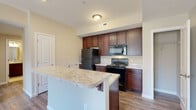 Located at 1564 McNaughten Rd in Columbus, Ohio, this single-floor residence offers a practical and comfortable layout designed for everyday living. The kitchen serves as a central highlight, showcasing rich dark-stained cabinetry that extends to the ceiling for ample storage, light granite-style countertops, and a full suite of black appliances including a refrigerator, electric range, over-the-range microwave, and dishwasher. A peninsula with a double-basin sink adds both workspace and a casual gathering point, while wood-look plank flooring runs throughout the kitchen and connecting areas, adding warmth and durability. Adjacent to the kitchen, a walk-in closet provides generous storage space. The living area offers a comfortable, open feel with neutral carpet, light-toned walls, and a sliding glass door that lets in natural light and provides access to a private balcony with outdoor views. The bathroom is finished with tile flooring in a neutral palette, featuring a full-size soaking tub and shower combination with a clean white surround. Throughout the space, a consistent neutral color scheme, textured ceilings, and well-placed ventilation contribute to a tidy, move-in-ready atmosphere suited for a variety of lifestyles.