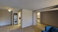 This single-floor residence offers a practical and comfortable layout designed for everyday living. The main living area features neutral-toned carpeting underfoot and a textured ceiling overhead, creating a cozy and understated ambiance. A central dividing wall helps define separate functional zones while maintaining an open feel throughout the space. Adjacent to the living area, the kitchen is outfitted with white cabinetry and tile flooring, providing a clean and bright workspace. A notable convenience of this home is the in-unit stacked washer and dryer, tucked neatly into a dedicated closet space just off the kitchen area, adding practicality without sacrificing space. The bathroom, visible from the main living area, features vanity lighting and white cabinetry, contributing to a bright and functional design. Throughout the residence, the neutral color palette of soft whites and warm grays creates a cohesive and inviting atmosphere. The single-floor layout ensures easy accessibility and seamless flow between spaces, making it a highly functional home suited for a range of lifestyles.