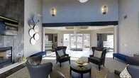 This well-appointed single-floor space presents a refined and welcoming atmosphere, centered around a spacious lounge area with dramatically high ceilings that create an open, airy feel. A bold dark structural beam serves as a striking architectural accent, contrasting elegantly against the soft gray walls and crisp white ceiling details. Recessed lighting and stylish wall sconces provide warm, layered illumination throughout.

The seating area is thoughtfully arranged with dark leather wingback chairs accented by decorative patterned pillows, surrounding a round gold-toned accent table — a combination that blends classic and contemporary design sensibilities. A navy blue bench adds a pop of color and additional seating near the periphery.

Large glass-paneled entry doors flood the interior with natural light, offering views of the surrounding outdoor landscape. Flanking the central lounge, open passageways lead to adjacent rooms or offices, suggesting a multi-functional layout suited for a communal or professional setting. Decorative circular wall art adds artistic character to one side of the room. The overall ambiance is polished and inviting, balancing sophisticated design elements with a comfortable, functional environment.
