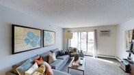 This single-floor residence centers around a welcoming living room that serves as the heart of the space. The room is finished with neutral gray walls and a textured ceiling that lends a classic character to the interior. Wall-to-wall carpeting in a warm beige tone adds comfort underfoot and contributes to the overall cozy atmosphere.

A standout feature of the living room is the large sliding glass door paired with an expansive window, allowing generous amounts of natural light to flood the space and offering views of the surrounding outdoor greenery. Sheer white curtains frame the opening, softening the light and adding an airy quality to the room. A wall-mounted air conditioning unit ensures year-round comfort.

The living area is well-proportioned, comfortably accommodating a full sofa arrangement, accent chairs, and a central coffee table. Bold, decorative artwork in shades of blue adorns the walls, providing a vibrant contrast to the otherwise understated color palette. A floor lamp positioned near the window supplements the natural light during evening hours, enhancing the room's warm and inviting ambiance.