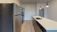 This single-floor unit showcases a contemporary design with a neutral gray and white color palette, light wood-look luxury vinyl plank flooring, and clean finishes throughout. The open-concept living and kitchen area serves as the heart of the space, where a teal-accented center island with a white quartz countertop and integrated sink takes center stage, illuminated by a sleek pendant light. The kitchen is equipped with stainless steel appliances, including a refrigerator, electric range, and over-the-range microwave, complemented by gray cabinetry, white countertops, and open wood shelving for additional storage. A sliding barn door adds a modern-industrial touch to the layout.

The living area is generously proportioned, featuring recessed and track lighting that brightens the neutral walls. A bedroom offers a window with exterior views, a built-in wardrobe, and consistent flooring. The bathroom is appointed with a long white vanity with ample drawer storage, a framed mirror, and a glass-enclosed walk-in shower. Throughout the unit, clean lines, modern fixtures, and a cohesive neutral aesthetic create a polished, move-in-ready environment.