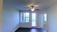 This single-story space features a clean, functional layout highlighted by a well-lit living area. The room is finished with dark hardwood-style flooring that contrasts neatly against the neutral, light gray walls, creating a balanced and modern aesthetic. A ceiling fan with an integrated light fixture is centrally positioned, providing both air circulation and ambient lighting throughout the space.

Natural light is a defining characteristic of this area, with a large window fitted with horizontal blinds allowing views of surrounding greenery. Adjacent to the window, a glass-paneled door with blinds opens directly onto what appears to be a private balcony, further enhancing the connection to the outdoors and bringing in additional natural light.

The room transitions smoothly into adjacent spaces, with a doorway visible to the right leading to additional areas of the unit. The overall ambiance is bright, clean, and move-in ready, with a straightforward design that offers a comfortable and practical living environment. The combination of natural light, outdoor access, and contemporary finishes gives the space an open, airy feel.