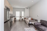 This single-floor studio is designed with a modern, minimalist aesthetic that maximizes functionality within an open-concept layout. The floor plan reveals a seamless flow between the sleeping, living, and kitchen areas, all finished with light wood-tone flooring that contributes to a bright and cohesive atmosphere throughout.

The kitchen is a standout feature, showcasing flat-front light wood cabinetry extending from floor to ceiling for generous storage. Stainless steel appliances include a range, over-the-range microwave, dishwasher, and refrigerator, complemented by grey quartz-style countertops and a modern undermount sink. A small breakfast bar provides casual seating.

The open living area accommodates both a seating arrangement and a sleeping space, with clean white walls and recessed lighting maintaining an airy, uncluttered feel. A wall-mounted mirror adds depth to the living zone.

The bathroom features a floating vanity with light wood cabinetry, a white countertop, small-format white mosaic tile flooring, and a clean, neutral color palette. Conveniently positioned adjacent to the bathroom is an in-unit washer, making laundry easily accessible. Overall, the space offers a practical and stylish layout suited for efficient single-floor living.