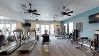 This fitness center offers a comprehensive workout environment thoughtfully designed to accommodate a range of exercise routines. The space is outfitted with an array of cardio equipment, including treadmills and elliptical machines, alongside a variety of strength training stations and cable machines, providing residents with everything needed for a well-rounded fitness regimen. The open floor plan allows for comfortable movement between equipment, with durable carpet-style flooring underfoot that adds both traction and comfort. Multiple ceiling fans are evenly distributed throughout the space, ensuring consistent airflow and a comfortable environment during workouts. Natural light streams in through several windows and a set of glass-panel doors, creating a bright and welcoming atmosphere. The walls feature a two-tone color scheme, with a calming teal accent wall complementing the neutral surrounding tones. A wall-mounted television adds an entertainment option for those looking to stay engaged during cardio sessions. Recessed lighting and a textured ceiling complete the practical yet inviting design of this well-maintained fitness facility.