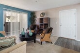 This model apartment at 18215 Foothill Blvd in Fontana, California, offers a well-designed single-floor layout with a variety of functional living spaces. The living room serves as the heart of the unit, featuring a bold teal accent wall, soft carpeting, and a large sliding glass door fitted with vertical blinds that opens to an outdoor patio, filling the space with natural light and providing a seamless indoor-outdoor connection. The kitchen is efficiently arranged with white cabinetry, a full suite of white appliances including a refrigerator and electric range, granite-style countertops, a double basin sink, and a skylight-style overhead fixture that brightens the workspace. Adjacent to the kitchen, the dining area includes a ceiling fan and offers a comfortable setting for everyday meals. The primary bedroom features a rich, dark accent wall, plush carpeting, and windows that allow in natural light. The second bedroom is carpeted as well, with a sliding closet and a vertical blind-covered window. Two bathrooms serve the unit — one with a tiled tub-shower combination and the other with a granite-style vanity countertop, white cabinetry, and tile flooring — both clean and functional in design.