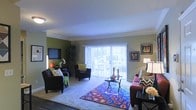 This single-story space centers around a spacious living room that combines comfort and style in a well-organized layout. The room is finished with plush wall-to-wall carpeting and features crown molding along the ceiling line, adding a refined architectural detail to the space. Walls are painted in a two-tone scheme, with an olive green accent wall providing a warm contrast to the neutral gray tones elsewhere.

Natural light flows generously into the living area through a large sliding glass door fitted with vertical blinds, offering a view of an outdoor patio area. The glass door serves as both a functional entry point and a key source of daylight, keeping the interior bright and airy.

The living room is thoughtfully arranged with a mix of seating, including a sofa and accent chairs, centered around a dark coffee table atop a vibrant, patterned area rug that adds a bold pop of color to the space. A wall-mounted flat-screen television and framed wall art complete the décor. A floor lamp and table lamp provide warm supplemental lighting, contributing to an inviting, comfortable ambiance throughout.