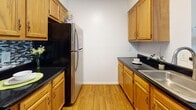 This single-floor unit presents a clean, functional layout designed for comfortable everyday living. The open living area is finished with neutral wall-to-wall carpeting and features a ceiling fan, creating a bright and airy atmosphere. A pass-through opening connects the living space to the kitchen, enhancing the sense of openness. The galley-style kitchen is well-appointed with warm honey-toned wood cabinetry, dark granite-style countertops, hardwood-style flooring, and stainless steel appliances including a full-size refrigerator. Ample upper and lower cabinet storage lines both sides of the kitchen, maximizing utility in the compact space. The bedroom is generously sized, featuring carpet flooring, a window that allows natural light to filter in, and direct access to additional rooms. A dedicated laundry area with bifold doors is conveniently located within the unit, adding practical value. The full bathroom includes a combination tub and shower with a clean white surround, hardwood-style flooring, a wood-toned vanity with dark countertop, and crisp white walls. Throughout the unit, the neutral color palette and consistent finishes create a cohesive and move-in-ready environment.