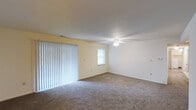This single-floor space centers around a spacious living area finished with wall-to-wall carpeting in a neutral gray-brown tone, providing a comfortable and cohesive foundation throughout the room. The walls are painted in a soft, light cream color, contributing to a clean and bright atmosphere. A ceiling fan with an integrated light fixture is centrally positioned, offering both overhead illumination and air circulation year-round. One wall features a large sliding glass door dressed with full-length vertical blinds, allowing for adjustable natural light and potential outdoor access. An additional window on the adjacent wall further enhances the natural light entering the space. Clean white baseboards run along the perimeter of the room, adding a crisp, finished detail to the interior. Multiple electrical outlets are positioned along the walls, offering practical functionality throughout the room. The overall layout is open and unobstructed, providing flexibility for a variety of furniture arrangements and uses. The space presents a straightforward, functional design with a neutral palette well-suited for personalization.