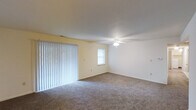 This single-floor space centers around a spacious living area finished with wall-to-wall carpeting in a neutral gray-brown tone, providing a comfortable and cohesive foundation throughout the room. The walls are painted in a soft, light cream color, contributing to a clean and bright atmosphere. A ceiling fan with an integrated light fixture is centrally positioned, offering both overhead illumination and air circulation year-round. One wall features a large sliding glass door dressed with full-length vertical blinds, allowing for adjustable natural light and potential outdoor access. An additional window on the adjacent wall further enhances the natural light entering the space. Clean white baseboards run along the perimeter of the room, adding a crisp, finished detail to the interior. Multiple electrical outlets are positioned along the walls, offering practical functionality throughout the room. The overall layout is open and unobstructed, providing flexibility for a variety of furniture arrangements and uses. The space presents a straightforward, functional design with a neutral palette well-suited for personalization.