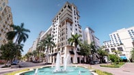 This residence is situated within an impressive multi-story building that showcases a Mediterranean-inspired architectural style, characterized by a light-toned stucco exterior, structured balconies with dark awnings, and clean geometric lines that rise prominently against a clear blue sky. The building's exterior is beautifully framed by tall, mature palm trees that lend a distinctly tropical character to the setting. At the front of the building, an ornamental fountain serves as a central focal point of the plaza, adding an element of elegance and vitality to the arrival experience. The ground level features retail or commercial frontage with signature dark canopy awnings, contributing to the mixed-use character of the development. Private balconies extend from each floor, offering residents outdoor space and views of the surrounding area. The overall ambiance of the building and its grounds reflects a well-maintained, upscale environment with thoughtful landscaping and architectural detailing throughout. The single-floor residence within this building benefits from the building's prominent positioning and refined communal spaces, making it part of a distinguished residential community defined by its graceful design and inviting atmosphere.