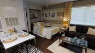 This single-floor studio-style space makes efficient use of its open layout by thoughtfully combining a sleeping area, dining space, and lounge zone into one cohesive environment. The design is anchored by a striking gray and yellow color palette, carried throughout via bold striped wall treatment, chevron-patterned curtains, and coordinating yellow accent pillows and bedding. Dark hardwood flooring runs throughout, grounding the space and adding warmth against the cooler gray tones. The sleeping area features a generously sized bed positioned against the accent wall, which is adorned with framed artwork and horizontal painted stripes, creating a focal point that adds visual depth. A pendant light overhead provides ambient illumination. Adjacent to the bed, a compact lounge area includes a dark upholstered armchair paired with a sleek coffee table topped with books and décor, offering a relaxed sitting space. A small dining table with chairs occupies the opposite corner, styled with a black-and-white geometric rug beneath. A window dressed with chevron drapes allows natural light to filter into the space, enhancing its bright and energetic character.