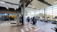 This fitness center is a thoughtfully designed, single-floor facility that combines functionality with a bright, energizing atmosphere. Soaring ceilings with exposed ductwork and structural columns lend the space an industrial-modern aesthetic, while a dramatic floor-to-ceiling glass curtain wall along one side fills the entire area with abundant natural light and open views of the surrounding greenery.

The facility is outfitted with a comprehensive range of True-brand cardio equipment, including treadmills and upright machines, arranged along the windowed wall to allow members to enjoy outdoor views during their workouts. A dedicated strength training zone features a multi-station cable and weight machine setup, complemented by a dumbbell rack and mirrored wall that enhances the sense of space while supporting proper form.

Underfoot, a stylish geometric-patterned vinyl flooring adds visual interest and provides durable, easy-to-maintain surface coverage throughout the workout areas. Linear pendant lighting supplements the natural light, ensuring the space remains well-lit at all hours. The overall ambiance is clean, modern, and motivating — a well-appointed fitness environment designed to support a full range of exercise routines in a spacious and comfortable setting.
