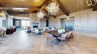 Located in Colorado Springs, CO, this single-floor community clubhouse offers a generously sized, thoughtfully designed gathering space that blends mountain-lodge aesthetics with modern sophistication. The main hall is anchored by soaring vaulted ceilings clad in warm wood planking and defined by bold dark-stained exposed timber beams. Dramatic antler-style chandeliers hang overhead, casting ambient light across a striking chevron-patterned tile floor. A stone fireplace serves as a focal point in the lounge area, complemented by upholstered seating arranged for comfortable social gatherings. Large windows throughout flood the interior with abundant natural light and frame sweeping views of the surrounding Colorado Front Range and Rocky Mountains. A full bar area features a stone-clad island counter with bar seating, wood-plank ceiling details, and wall-mounted screens. A dedicated game room offers a billiards table set on plaid-patterned carpet beneath a tray ceiling with a statement light fixture. Throughout the space, warm-toned wood paneling, neutral furnishings, and layered textures create an inviting, resort-inspired ambiance well suited for both community events and casual everyday use.