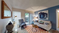 This single-floor residence offers a thoughtfully designed layout that balances comfort and functionality. The living area serves as the centerpiece of the space, featuring plush carpeting, a bold chevron-patterned area rug, and a striking blue accent wall adorned with large-scale artwork. A sliding glass door with vertical blinds opens directly onto a private patio, allowing natural light to fill the room and providing pleasant outdoor views of surrounding greenery. A console table with a mirror adds a decorative focal point near the entryway transition.

The kitchen is efficiently laid out with rich dark cabinetry, white countertops, and black appliances including a refrigerator, electric range, and over-the-range microwave. Warm hardwood-style flooring runs through the kitchen, contrasting nicely with the carpeted living and sleeping areas.

The bedroom is a comfortable retreat, highlighted by a teal accent wall, ceiling fan with light fixture, and a window overlooking the outdoors. Carpeted flooring and ample wall space contribute to a calm, restful atmosphere.

The bathroom features a full tub-shower combination with white tile surround, a vanity with stone-topped counters, and hardwood-style flooring, completing this well-appointed residence.