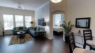 This single-story space showcases a thoughtfully designed open-concept layout with rich hardwood flooring flowing throughout. The living area is anchored by a pendant light fixture and a ceiling fan, both contributing to a well-lit and comfortable atmosphere. Crown molding lines the upper perimeter of the walls, adding a refined architectural detail that elevates the overall aesthetic. Large windows fitted with horizontal blinds allow generous natural light to fill the living space, which features a coordinated seating arrangement including a sofa and armchair in cool blue-gray tones, complemented by decorative throw pillows. A round coffee table and end tables complete the sitting area, which is grounded by a patterned area rug over the hardwood floor. An elegant arched passageway connects the living area to the bedroom, lending an architectural focal point to the space. The bedroom, visible through the archway, is bright and serene, with a neatly made bed, a bedside lamp, and a small plant adding a touch of warmth. A tall indoor palm plant near the dining area introduces a natural, tropical element to the space's overall ambiance.