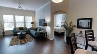 This single-story space showcases a thoughtfully designed open-concept layout with rich hardwood flooring flowing throughout. The living area is anchored by a pendant light fixture and a ceiling fan, both contributing to a well-lit and comfortable atmosphere. Crown molding lines the upper perimeter of the walls, adding a refined architectural detail that elevates the overall aesthetic. Large windows fitted with horizontal blinds allow generous natural light to fill the living space, which features a coordinated seating arrangement including a sofa and armchair in cool blue-gray tones, complemented by decorative throw pillows. A round coffee table and end tables complete the sitting area, which is grounded by a patterned area rug over the hardwood floor. An elegant arched passageway connects the living area to the bedroom, lending an architectural focal point to the space. The bedroom, visible through the archway, is bright and serene, with a neatly made bed, a bedside lamp, and a small plant adding a touch of warmth. A tall indoor palm plant near the dining area introduces a natural, tropical element to the space's overall ambiance.