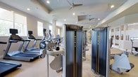 This fitness center offers a comprehensive workout environment designed to accommodate a range of exercise needs. The space is outfitted with Matrix-brand strength training machines positioned prominently throughout the floor, providing users with a variety of resistance and muscle-targeting options. Alongside the strength equipment, a row of cardio machines — including treadmills and elliptical trainers — lines one side of the room, offering ample options for aerobic exercise.

The interior features a clean, neutral color palette with light-toned walls and flooring that contributes to an open, airy atmosphere. Tray ceiling details add architectural interest and a sense of vertical space, while recessed lighting ensures the area is well-illuminated throughout. Ceiling fans are installed overhead, promoting air circulation and comfort during workouts. Natural light filters in through windows along the wall, further brightening the environment.

Large mirrors are strategically placed to visually expand the space and allow users to monitor their form during exercise. The overall layout is organized and spacious, providing comfortable movement between equipment stations and creating a functional, inviting fitness environment.