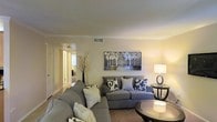 This single-floor residence offers a thoughtfully designed layout that balances comfort and style. The living room serves as the heart of the space, showcasing warm neutral wall tones that create an inviting and relaxed atmosphere. A plush gray sectional sofa, accented with patterned throw pillows in black and white, anchors the room and provides generous seating. A round glass-top coffee table and a sleek side table with a contemporary lamp add functional elegance to the space. A framed landscape painting of white trees hangs prominently above the sofa, serving as a striking focal point and adding artistic character to the room. Crown molding along the ceiling lends a refined architectural detail, while recessed vents indicate a central air system for year-round comfort. A decorative arrangement of branches in a tall vase introduces a touch of organic texture near the corridor. The hallway visible from the living area leads further into the residence, hinting at additional rooms beyond. Neutral-toned flooring flows consistently throughout, tying the spaces together seamlessly. Overall, the space exudes a calm, well-curated ambiance suited for comfortable everyday living.