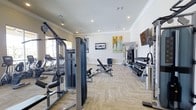 This fitness center offers a comprehensive and thoughtfully arranged workout environment designed to support a wide range of exercise routines. The space is finished with light herringbone-patterned flooring that lends a clean, modern aesthetic throughout. Recessed lighting in a tray ceiling provides bright, even illumination across the entire area.

The strength training section is anchored by a Matrix cable machine system, complemented by an adjustable weight bench and a fully stocked dumbbell rack with a broad range of free weights. TRX suspension training straps are mounted along one wall, offering versatile bodyweight training options. Cardio equipment, including treadmills, is positioned near the windows, allowing natural light to enhance the workout experience.

Wall-mounted flat-screen televisions are positioned for easy viewing from multiple areas of the floor, making workouts more engaging. Colorful artwork adorns the walls, adding a welcoming and vibrant touch to the otherwise clean, neutral interior palette. The overall layout is open and well-organized, ensuring easy movement between equipment stations. This fitness center provides residents or users with a fully functional facility suited for both strength and cardiovascular training needs.