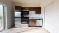 This single-floor space offers a clean, contemporary design with a cohesive aesthetic throughout. The kitchen is a standout feature, showcasing rich dark-walnut cabinetry that extends to the ceiling, providing ample storage above and below. Granite countertops add a touch of elegance, while a full suite of stainless steel appliances — including a refrigerator, electric range, over-the-range microwave, and dishwasher — ensures a fully functional cooking space. Light-toned luxury vinyl plank flooring in the kitchen creates a bright, welcoming atmosphere.

The living area is open and versatile, bathed in natural light streaming through a large sliding glass door that opens to an outdoor view of surrounding trees and the streetscape beyond. The warm-toned hardwood-style flooring in this area contrasts beautifully with the neutral, light-gray walls, creating a calm and inviting environment.

The bathroom is thoughtfully designed with matching hardwood-style flooring, a pedestal sink, and a well-placed mirror that enhances the sense of space. Clean white walls and efficient lighting give the bathroom a crisp, utilitarian feel. Throughout the unit, consistent finishes and neutral tones create a cohesive, modern living environment ready to be personalized.
