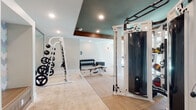 This fitness center is thoughtfully designed to support a comprehensive workout experience within a spacious, single-level layout. The interior is anchored by a striking tray ceiling finished in teal and framed by rich wood beam accents, creating a polished and modern aesthetic. Recessed lighting throughout ensures the space is consistently well-lit, complemented by an abundance of natural light streaming in through large glass panel doors and windows that offer views to an outdoor area.

The flooring features a patterned carpet in neutral tones, providing comfort underfoot while maintaining a clean, structured appearance. Support columns are integrated thoughtfully into the layout, helping to define distinct functional zones without disrupting the open flow of the space.

The strength training area is well-stocked, featuring a cable machine, a weight rack with an extensive plate collection, a dumbbell rack with a range of free weights, and multiple adjustable benches. A mirrored wall in the strength zone enhances the sense of space and aids in form monitoring. The cardio section houses several treadmills, an elliptical, and additional cardio equipment positioned near the natural light sources. A wall-mounted television is also present, adding an entertainment option for users. A ceiling fan provides additional air circulation throughout the room.