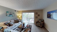 This single-floor residence offers a welcoming and well-appointed living space designed for comfortable everyday living. The living room serves as the heart of the home, featuring plush carpeting, a textured ceiling, and a large sliding glass door framed by vertical blinds that floods the room with natural light. The sliding door opens directly onto a private balcony, providing a seamless connection to the outdoors and offering pleasant views of the surrounding greenery. The living area comfortably accommodates a full seating arrangement, including a sofa, accent chair with a bold floral pattern, and a central coffee table set atop a decorative area rug, creating a stylish yet functional gathering space. Warm wall tones complement the neutral carpeting throughout, contributing to an inviting ambiance. A wall-mounted air vent ensures year-round climate comfort. Decorative wall art adds personal character to the space, while a table lamp provides warm supplemental lighting for evenings. The balcony, visible through the glass door, offers outdoor seating and serves as a private retreat. Overall, this residence combines practical features with a comfortable, lived-in aesthetic suited for relaxed daily living.