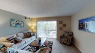 This single-floor residence offers a welcoming and well-appointed living space designed for comfortable everyday living. The living room serves as the heart of the home, featuring plush carpeting, a textured ceiling, and a large sliding glass door framed by vertical blinds that floods the room with natural light. The sliding door opens directly onto a private balcony, providing a seamless connection to the outdoors and offering pleasant views of the surrounding greenery. The living area comfortably accommodates a full seating arrangement, including a sofa, accent chair with a bold floral pattern, and a central coffee table set atop a decorative area rug, creating a stylish yet functional gathering space. Warm wall tones complement the neutral carpeting throughout, contributing to an inviting ambiance. A wall-mounted air vent ensures year-round climate comfort. Decorative wall art adds personal character to the space, while a table lamp provides warm supplemental lighting for evenings. The balcony, visible through the glass door, offers outdoor seating and serves as a private retreat. Overall, this residence combines practical features with a comfortable, lived-in aesthetic suited for relaxed daily living.