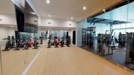 This contemporary fitness facility is thoughtfully designed to accommodate a wide range of workout routines within a clean, modern environment. The space is laid out across a single level, maximizing accessibility and ease of movement throughout.

The main workout area features light wood-toned flooring that lends a bright, welcoming atmosphere, complemented by crisp white walls and recessed lighting that ensures even illumination throughout. Full-length mirrors line one wall, creating a sense of expanded space while allowing users to monitor their form during exercise.

A dedicated cycling area is outfitted with a row of stationary spin bikes alongside stability and medicine balls, making it well-suited for both individual training and group cycling classes. Adjacent to this zone, a glass-enclosed strength training room houses a comprehensive selection of resistance machines and cable systems, offering a visually open yet acoustically separated environment for weight training.

Ventilation is integrated into the ceiling, supporting comfort during high-intensity workouts. The overall design strikes a balance between functionality and aesthetics, with clean lines, an uncluttered layout, and generous open floor space that accommodates both structured classes and independent training sessions. The facility presents a polished, professional atmosphere suitable for a variety of fitness levels and disciplines.