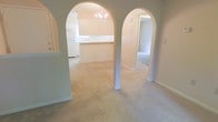 This single-floor residence showcases a thoughtfully designed interior defined by its most distinctive architectural feature: a pair of graceful arched doorways that frame transitions between the main living area and the adjoining spaces. The arches lend the home a classic, refined character and create a natural sense of flow throughout the layout.

The living area is finished with wall-to-wall neutral carpeting that adds warmth and comfort underfoot. Soft, warm-toned walls throughout the home contribute to a bright and welcoming ambiance. Through one of the arched openings, the kitchen comes into view, featuring a raised breakfast bar or counter that provides both a functional workspace and a casual dining option. White appliances, including a refrigerator, are visible, and the space benefits from natural light streaming through a window above the sink area.

A chandelier is suspended within the kitchen or dining area, adding an elegant decorative touch that elevates the overall aesthetic. The second arched doorway leads to a carpeted hallway or additional room, suggesting a well-organized and spacious floor plan. Clean white trim and baseboards frame each room, providing crisp contrast against the warm wall tones. Overall, the space offers a combination of classic architectural detail and practical, comfortable living.