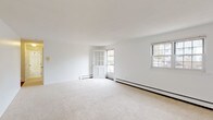 This single-floor, one-bedroom, one-bathroom unit offers a clean and functional layout designed for comfortable everyday living. The living area is generously sized, featuring neutral wall-to-wall carpeting, crisp white walls, and multiple windows that invite abundant natural light. Baseboard heating runs along the perimeter, ensuring warmth throughout. A charming glass-paneled door leads to an outdoor access point, enhancing the sense of openness. The kitchen is well-appointed with white cabinetry featuring wood-trimmed accents, dark granite-style countertops, and hardwood flooring. It includes a full suite of appliances — a refrigerator, electric range, dishwasher, and sink — making it a practical and efficient workspace. The bedroom is a comfortable, carpeted retreat with a large window overlooking the outdoors and a built-in closet with a rod for ample storage. It connects conveniently to the kitchen area. The bathroom is finished with a classic black-and-white octagonal tile floor, a white vanity with storage, a toilet, and a tub-shower combination enclosed by a white curtain — all presenting a timeless, clean aesthetic throughout.