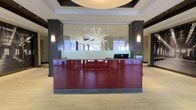 This sophisticated lobby space makes an immediate impact with its carefully considered design and high-end finishes. At the center of the space stands a commanding reception desk finished in deep burgundy lacquer with a granite countertop, flanked by dark charcoal-toned structural columns that add a sense of gravitas and definition to the area. Behind the desk, a frosted glass panel partition provides both privacy and an elegant backdrop, reflecting a sculptural starburst chandelier overhead that serves as a dramatic focal point.

The ceiling features a recessed circular tray detail that frames the chandelier and adds architectural depth, while recessed lighting throughout ensures the space is well-illuminated. Wall-mounted sconces on either side of the reception area contribute a warm, layered lighting effect. Large windows allow natural light to filter through, enhancing the bright and open ambiance of the lobby.

The flooring is composed of large-format neutral tiles, lending a clean and expansive feel to the overall space. Subtle greenery is introduced through potted plants near the desk, softening the structured design. The overall aesthetic is contemporary and professional, conveying a polished, corporate environment well-suited for a hotel, office building, or conference center.