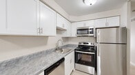 This single-floor residence offers a thoughtfully designed layout that balances comfort and functionality throughout its living spaces. The kitchen is a standout feature, outfitted with crisp white shaker-style cabinetry, marble-patterned countertops, and a full suite of stainless steel appliances including a refrigerator, electric range, over-the-range microwave, and dishwasher. Light wood-look flooring runs through the main living areas, lending a clean and contemporary aesthetic. The spacious living room is anchored by a large sliding glass door that fills the room with natural light and provides direct access to the outdoors, offering views of surrounding greenery. A built-in shelving nook adds a practical and decorative element to the living space. The bedrooms are finished with plush carpeting and feature generous closet storage with louvered bifold doors and built-in shelving. The bathroom is finished in a bright, neutral palette with diamond-pattern white tile in the tub surround, a full soaking tub, and light flooring that complements the overall design. Crown molding and clean white trim details are carried throughout, contributing to a polished and cohesive interior character.