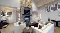 This single-floor space makes an immediate impression with its soaring ceilings and a striking floor-to-ceiling stone fireplace that serves as the undeniable focal point of the main lounge area. The fireplace, crafted from natural-toned stacked stone, features an elegant arched recess housing a wall-mounted flat-screen television above a classic dark mantel, creating a seamless blend of rustic and contemporary design.

The lounge area is thoughtfully arranged with plush white seating, including sofas and accent chairs accented by earth-toned throw pillows, surrounding a dark espresso coffee table. A hexagonal wooden side table and a potted plant add organic warmth to the setting. The light hardwood flooring complements the neutral palette of warm beige and grey tones throughout the space.

Natural light filters in through windows and a glass-paned door, brightening the interior and enhancing the airy feel created by the generous ceiling height. Subtle recessed lighting and ceiling speakers suggest a space designed for both relaxed gatherings and functional use. Additional rooms visible through doorways hint at further amenities beyond the main lounge, making this a well-appointed and inviting communal environment.