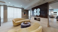 Located at 25 William T Morrissey Blvd in Boston, MA, this refined single-floor space presents a thoughtfully designed communal environment that blends contemporary architecture with curated artistic elements. The layout flows seamlessly across several distinct zones, each offering its own character while maintaining a cohesive aesthetic.

A curved wall of floor-to-ceiling sheer drapery defines one lounge area, flooding the space with soft, diffused natural light and framing bold orange accent chairs alongside a sleek dark wood reception desk. Structural columns with a brushed metallic finish punctuate the open floor plan, while large glass-panel partitions create transparency between zones without sacrificing definition.

An art gallery alcove features tiered, backlit display shelves showcasing rows of colorful framed works, complemented by teal swivel chairs and natural wood stump side tables—offering an intimate setting for quiet reflection. A dedicated lounge area centers around a linear fireplace set against a rich dark tile surround, with sculptural wall art above and curved, plush seating in warm golden tones arranged around a velvet ottoman.

Throughout the space, recessed lighting, neutral carpet flooring, and a muted gray-and-taupe color palette create a calm, upscale ambiance. The overall design communicates a professional yet welcoming environment, ideal for gathering, relaxation, or creative inspiration.