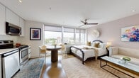This thoughtfully designed studio unit occupies an elevated floor, offering sweeping panoramic views of the surrounding cityscape through a generous bank of floor-to-ceiling windows. Natural light floods the open-plan living space throughout the day, complemented by warm hardwood flooring and a neutral, soft-toned color palette that creates a calm and inviting ambiance.

The main living area seamlessly integrates sleeping, dining, and lounging zones. A queen-sized bed with an arched headboard serves as the central anchor, flanked by layered area rugs and accented by a decorative juju hat wall piece. A ceiling fan overhead adds both style and function. A compact round dining table with wicker chairs sits near the windows, making the most of the elevated views. A loveseat and glass-topped side table complete the seating arrangement.

The kitchen is efficiently designed with sleek white cabinetry, white countertops, and a full suite of stainless steel appliances, including a range, microwave, dishwasher, and refrigerator. A closet provides additional storage and organization.

The bathroom is finished with large-format stone-look tile, a soaking tub, a floating vanity with an illuminated mirror, and clean, modern fixtures throughout — delivering a polished, spa-like feel.