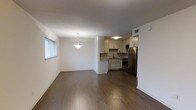 This single-floor space showcases an open-concept design that seamlessly connects the living and dining areas to the kitchen. The living and dining space is finished with rich, dark hardwood flooring that extends throughout, complemented by clean white walls and white baseboard trim that lend a bright, airy feel. A simple pendant light fixture hangs above the dining area, adding a subtle decorative touch. Natural light filters in through a horizontal blind-covered window along one wall.

The kitchen is efficiently laid out in an L-shaped configuration, featuring white cabinetry with ample upper and lower storage, a neutral tile backsplash, and a countertop with an integrated sink. Stainless steel appliances — including a refrigerator, range, and microwave — provide a modern contrast to the white cabinetry. The kitchen flooring continues the hardwood theme seen in the main living area, maintaining visual continuity across the space. A ceiling-mounted light fixture illuminates the kitchen, and a visible HVAC vent ensures year-round climate comfort. The overall ambiance is clean, functional, and move-in ready.