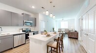 This single-floor residence offers four bedrooms, two bathrooms, and a thoughtfully arranged layout that maximizes functionality. The open-concept kitchen is a standout feature, equipped with stainless steel appliances including a refrigerator, electric range, and microwave, all complemented by sleek gray cabinetry and crisp white countertops. A central island with pendant lighting overhead provides additional prep space and casual seating, flowing naturally into the combined dining and living area. The living room is bright and welcoming, featuring light wood-style flooring, a ceiling fan, and windows that invite natural light. Soft neutral tones carry throughout the home, creating a clean and cohesive aesthetic. The bedrooms are carpeted for comfort, with the primary bedroom offering direct outdoor access through a glass door and windows with blinds for natural light and privacy. Two walk-in closets with wire shelving provide ample storage. The bathrooms are finished with gray vanities, white countertops, wood-style flooring, and tub-shower combinations. Recessed lighting and panel-style doors add to the modern, clean character of the home.