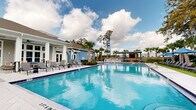 This community outdoor amenity space centers around a generously sized resort-style swimming pool with striking cobalt blue tile accents and clearly marked depth indicators. The crystal-clear turquoise water is complemented by a wide surrounding deck that provides ample space for lounging and socializing. A covered pavilion with a teal-accented roof serves as an outdoor entertainment hub, equipped with a full grilling station and seating area — ideal for gatherings. Additional poolside amenities include rows of chaise lounge chairs and shaded umbrella seating stations positioned along the perimeter, offering both sun and shade options for residents. The pool area is framed by mature palm trees and lush greenery, lending a tropical ambiance to the setting. A charming blue and white clubhouse building anchors one end of the pool deck, featuring large windows, white columns, and multiple access points that blend seamlessly with the outdoor environment. The surrounding landscape includes mature oak and pine trees that provide a natural canopy backdrop. Under open skies with abundant natural light, this thoughtfully designed outdoor space delivers a welcoming, vacation-inspired atmosphere suited for relaxation and community recreation year-round.
