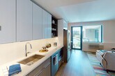 This single-floor studio is thoughtfully designed with a contemporary open-concept layout that maximizes functionality and flow. The kitchen runs along one wall in a galley-style configuration, outfitted with high-gloss light gray upper cabinetry, matte lower cabinets, and crisp white quartz countertops. Stainless steel appliances include a French door refrigerator, a slide-in range with a stainless wall-mounted range hood, a built-in microwave, and a dishwasher. Open shelving provides additional display and storage space, while under-cabinet lighting adds a warm, polished finish. A single-basin undermount sink with a modern arc faucet completes the kitchen.

The living and sleeping area occupies the main open space, finished with light wood flooring throughout. Natural light pours in through a teal-framed glass door that opens onto a private balcony, offering views of the surrounding cityscape and waterfront. The space accommodates both a seating area and a full bed, with neutral walls providing a versatile backdrop.

The bathroom features a glass-enclosed walk-in shower with floor-to-ceiling tile, a floating white vanity with an integrated sink, and a backlit mirror cabinet, creating a clean, spa-like atmosphere.