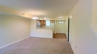 This two-floor residence offers a welcoming, open-concept layout on the main level, where the living area and kitchen flow seamlessly into one another. The living space is finished with plush neutral carpeting, contributing to a warm and comfortable atmosphere, while the kitchen and entry areas feature hard surface flooring that provides a practical contrast.

The kitchen is equipped with light wood cabinetry and includes a built-in microwave, refrigerator, and ample counter space along a peninsula that doubles as a breakfast bar or workspace. A window above the kitchen sink allows natural light to filter into the space. Overhead, a flush-mount ceiling light keeps the kitchen well-illuminated, while an elegant chandelier serves as a focal point above the adjoining living area.

The entryway, visible from the main living space, features a paneled door with decorative glass inserts that allow additional natural light into the home. A hallway leads to additional rooms, with multiple doors suggesting well-separated functional zones. Ventilation is provided throughout via ceiling-mounted HVAC registers. The overall aesthetic is clean, neutral, and move-in ready, with a straightforward design that suits a variety of lifestyles.