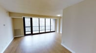 This single-floor unit offers a spacious, open-concept living area characterized by clean lines and a modern, minimalist aesthetic. The most prominent feature is the expansive wall of floor-to-ceiling windows paired with wide sliding glass doors, which flood the interior with natural light and provide direct access to an outdoor balcony. The views through the glass reveal a vibrant urban setting with neighboring high-rise buildings, reinforcing the elevated city-living atmosphere. The flooring throughout is smooth light-toned hardwood, lending warmth and continuity to the space. Walls are finished in a neutral cream tone, creating a versatile backdrop suitable for a range of interior styles. White baseboards provide a crisp contrast along the perimeter. A flush-mounted ceiling light offers functional ambient illumination. The room is generously proportioned, allowing for a variety of furniture arrangements. The overall ambiance is bright, airy, and contemporary, with the abundance of natural light and the seamless indoor-outdoor connection serving as key defining characteristics of this well-appointed urban living space.