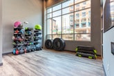 This single-level fitness facility in Atlanta, GA offers a thoughtfully designed workout environment tailored for functional and strength training. The space features durable wood-look flooring throughout, providing a clean, modern aesthetic that complements the cool gray walls and contemporary finishes.

The equipment zone is a standout area, housing two fully stocked vertical storage racks loaded with medicine balls, slam balls, sandbags, stability balls, and kettlebells in various weights, catering to a wide range of training styles. Large truck tires positioned nearby add a functional training element for more intensive workouts. Aerobic step platforms are also available and conveniently stored near the perimeter.

Large floor-to-ceiling windows line portions of the space, flooding the interior with natural light and offering views of the surrounding Atlanta streetscape. Glass-paneled partitions and doors divide sections of the facility, creating defined workout zones while maintaining an open, connected feel. A wall-mounted television and framed artwork contribute to an engaging atmosphere.

The overall design balances functionality and accessibility, making this facility a comprehensive destination for varied fitness routines.