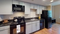 This single-floor studio unit offers a practical and well-organized layout that combines living and sleeping areas within one open space. The main room features warm hardwood-style flooring, neutral wall tones, and a window fitted with blinds that allows natural light to brighten the interior. The space comfortably accommodates both a seating area and a sleeping area, providing functional flexibility.

The kitchen is efficiently designed with a single-wall configuration, featuring gray cabinetry extending to ceiling height for generous storage. Dark granite-style countertops contrast with the light cabinetry, and black appliances — including a range, microwave, dishwasher, and refrigerator — complete the setup. The hardwood-style flooring continues seamlessly from the main living area into the kitchen, reinforcing a cohesive aesthetic throughout.

The bathroom is accessed through a hallway and features large-format dark tile flooring, providing a grounded contrast to the light-colored walls. A shower with a decorative curtain and an adjacent walk-in closet with a hanging rod offer practical storage and convenience. Recessed lighting and a clean, minimal design give the bathroom a tidy, functional character. Overall, the unit presents a straightforward, well-maintained living environment.