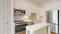 This thoughtfully designed one-bedroom, one-bathroom unit offers a clean, modern aesthetic throughout its well-appointed single floor. The open-concept kitchen is a standout feature, showcasing crisp white shaker cabinetry, a textured tile backsplash, gleaming quartz countertops, and a functional center island. A full suite of stainless steel appliances — including a range, over-the-range microwave, dishwasher, and refrigerator — rounds out the kitchen's practical appeal. Light hardwood-style flooring flows seamlessly through the main living areas, enhancing the bright, airy atmosphere. A hallway with a barn-style door connects the kitchen to the living room, which features a ceiling fan and large sliding glass doors that open to an outdoor space, flooding the room with natural light and offering open exterior views. The generously sized primary bedroom is finished with soft carpeting, a ceiling fan, and convenient access to a dedicated closet. An in-unit laundry area with a stacked washer and dryer adds everyday convenience. The overall design palette of whites and warm neutrals creates a cohesive, modern living environment that feels both functional and inviting.