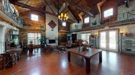 This expansive single-floor space is defined by its dramatic rustic-Western character, showcasing soaring cathedral ceilings with exposed timber beams and wood-plank decking that create an immediate sense of grandeur. The open great room serves as the central hub of the space, anchored by a floor-to-ceiling stacked stone fireplace flanked by leather seating and accented with a decorative tin wall piece. Antler and wrought-iron chandeliers cast warm ambient light throughout, complementing the wall sconces positioned around the perimeter. Polished stained concrete floors in deep reddish-brown tones run throughout, contributing to the rugged aesthetic. An upper mezzanine level, accessible via a rustic staircase and enclosed by a branch-crafted railing, overlooks the main floor below. Unique hand-carved wood furnishings—including a sculptural live-edge bench and a wagon wheel display piece—reinforce the Western theme. A dedicated seating area with a wall-mounted television offers a media-focused retreat, while a checkers table set near large windows with natural light adds a casual, social element. Double glass entry doors open to the exterior, inviting natural light and outdoor views into the space.