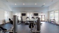 Located at 145 Retreat Dr in Pittsboro, NC, this single-floor fitness center is a well-appointed facility designed to support a wide range of workout routines. The space is bright and open, featuring white walls, dark rubberized flooring, and a tray ceiling with recessed lighting that creates a clean, modern aesthetic throughout.

The cardio zone is lined with multiple treadmills and an elliptical trainer, positioned in front of large windows that allow natural light to flow in and offer views to the outdoors. Wall-mounted flat-screen TVs are positioned throughout this area for entertainment during workouts. French doors with glass panels provide additional natural light and direct access to the exterior.

The strength training area is generously equipped with a variety of True-brand weight machines, a cable system, free weights including dumbbells and kettlebells, and adjustable benches. A dedicated functional training zone features suspension trainers (TRX), wall-mounted rig systems, medicine balls, stability balls, and organized storage cubbies. Full-length mirrors along one wall enhance the sense of space and assist with form during exercise.

The layout allows for ample circulation between equipment stations, making the facility practical and accessible for multiple users simultaneously. Overall, the fitness center offers a comprehensive and well-organized environment suitable for cardiovascular, strength, and functional training needs.