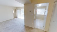 This single-floor residence features an open and functional layout designed to maximize usable living space. The main living area is finished with neutral-toned carpeting that adds warmth underfoot, while large sliding glass doors with vertical blinds allow an abundance of natural light to fill the room and provide access to an outdoor area. A front entry door is also visible, offering a welcoming point of arrival.

A central dividing wall separates the living space from an adjoining room, which transitions into tile flooring — indicative of a kitchen or utility area. This secondary space benefits from its own natural light source through a window dressed with vertical blinds, and features overhead lighting for added brightness. A fire extinguisher is mounted on the wall, reflecting attention to safety.

Throughout the unit, the walls are finished in a clean, neutral cream tone that complements both the carpeted and tiled flooring. The overall ambiance is bright, airy, and move-in ready, with a straightforward layout that lends itself well to a variety of furniture arrangements and living configurations.