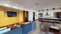 This single-floor space showcases a thoughtfully designed open-concept layout that effortlessly connects the living, dining, and kitchen areas into one cohesive environment. Light hardwood flooring runs throughout, providing a warm and inviting foundation that complements the clean white walls and modern recessed lighting overhead.

The living area is anchored by a plush blue sectional sofa accented with colorful throw pillows, positioned across from a wall-mounted flat-screen television set against a warm yellow accent wall. Rich dark wood cabinetry flanking the TV adds depth and a touch of sophistication to the space.

Transitioning seamlessly from the living area, the dining space features a sleek bar-height table paired with wire-frame chairs, offering a contemporary and casual setting for meals or gatherings. A decorative deer head wall mount adds a subtle rustic charm to the overall modern aesthetic.

The kitchen is well-appointed with dark wood upper cabinetry, a stainless steel refrigerator, and a stylish tiled backsplash in muted gray tones. Pendant lighting hangs above the counter, providing both functional illumination and an elegant design detail. The open flow between all areas makes this space highly functional, social, and visually expansive.